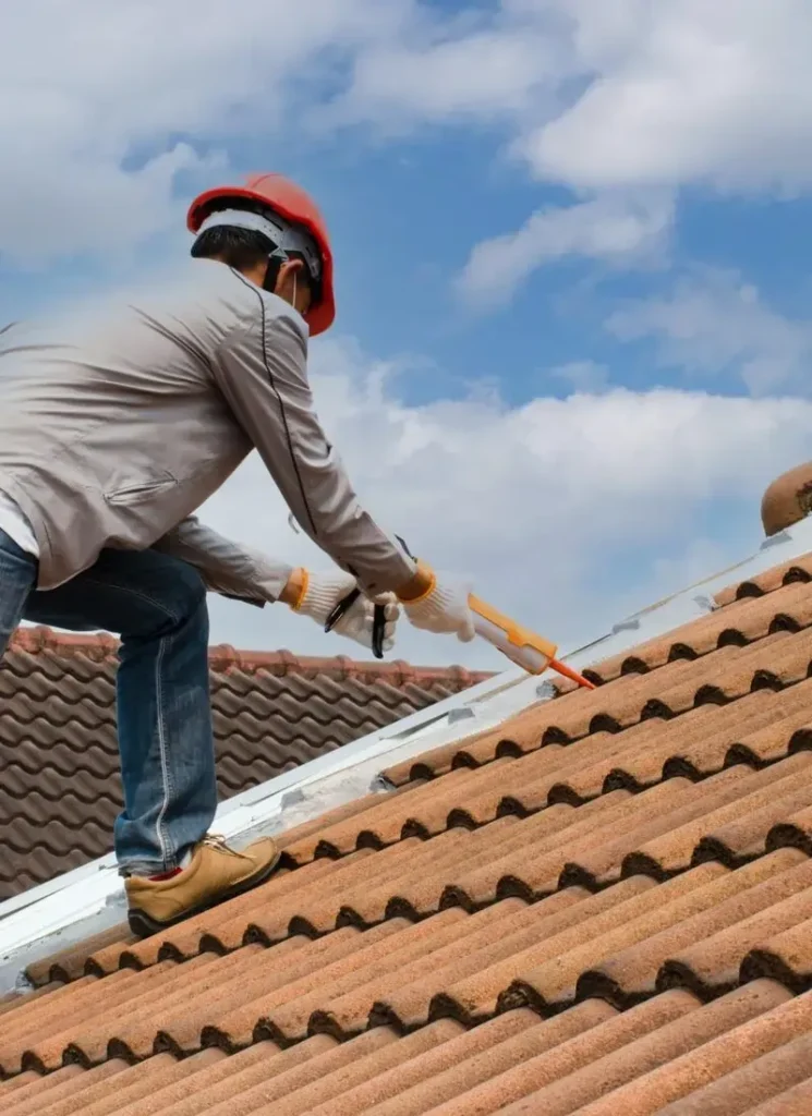 Home A construction worker in a safety helmet applies sealant to a tiled roof under a blue sky with scattered clouds.