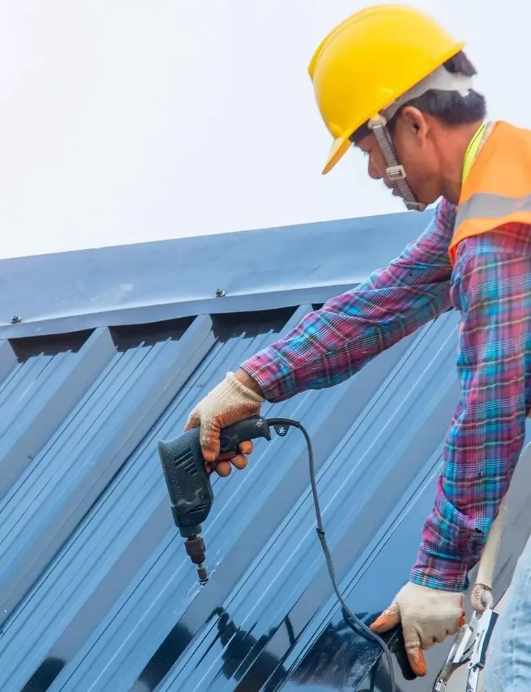 Home A worker in a yellow hard hat and gloves uses a drill to secure metal roofing