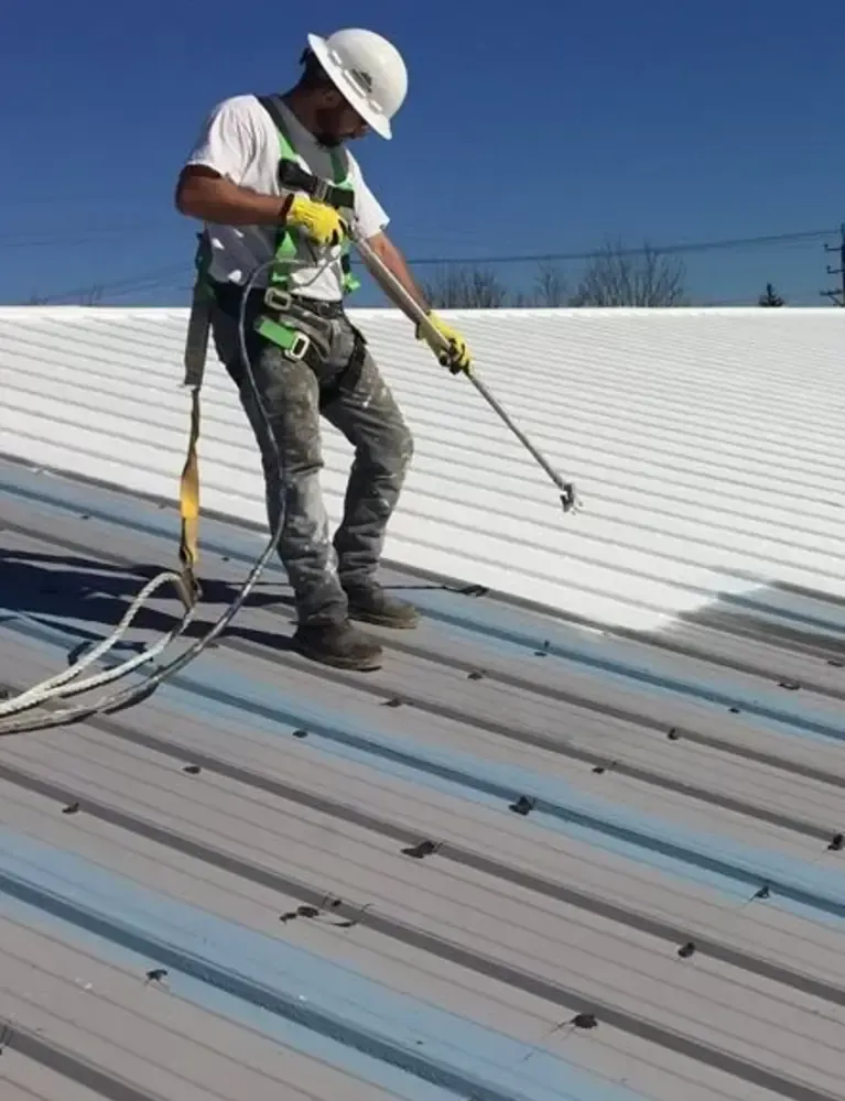 Home A worker in safety gear uses a roller to apply a white coating on a metal roof