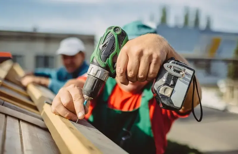 A worker holds a power drill, fastening wooden planks on a roof